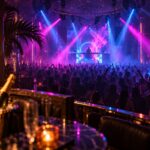 A packed Miami nightclub dance floor lit in magenta and blue beams, viewed from a VIP table toward the DJ stage